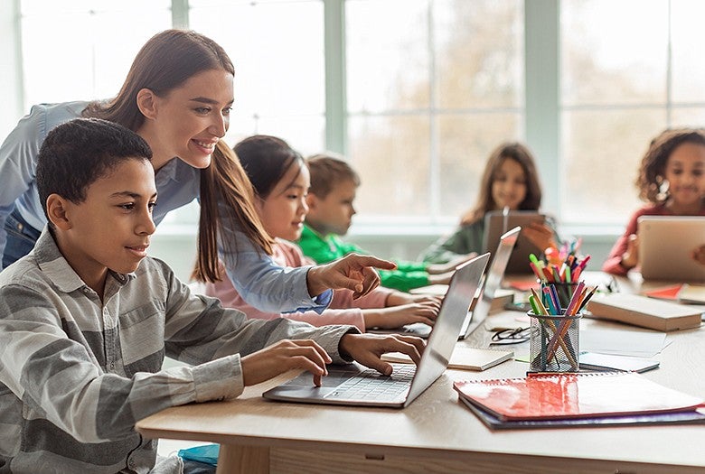 children sitting in class with teacher while learning on laptops