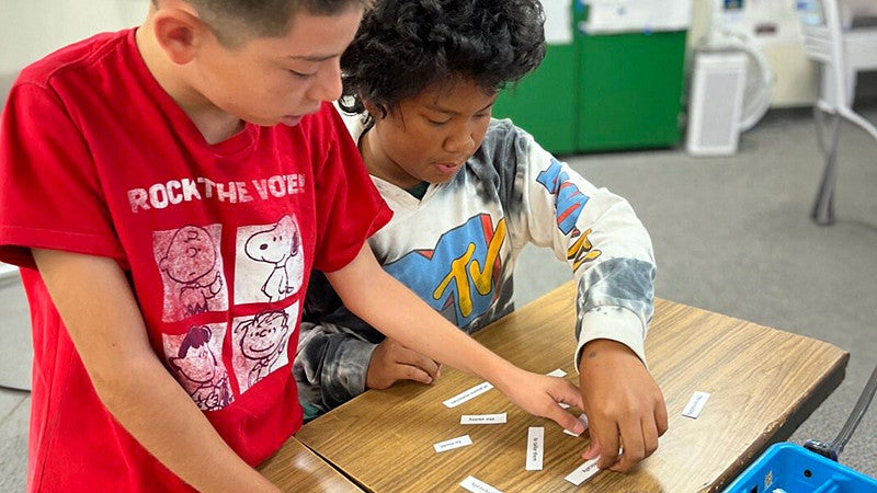 two young individuals working on a word puzzle on top of a classroom table