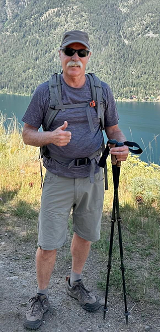Bruce Waltz standing in an outdoor setting in hiking gear giving a thumbs up sign