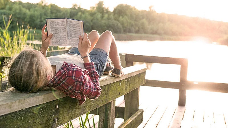 child laying on a bench in front of a lake while reading a book