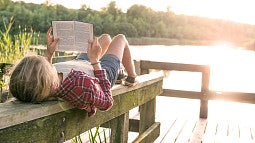 child laying on a bench in front of a lake while reading a book
